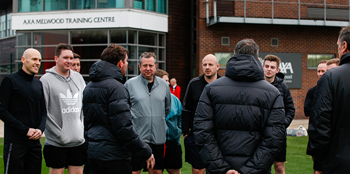 Group of people stood outside AXA Melwood Training Centre next to the sign with coach behind